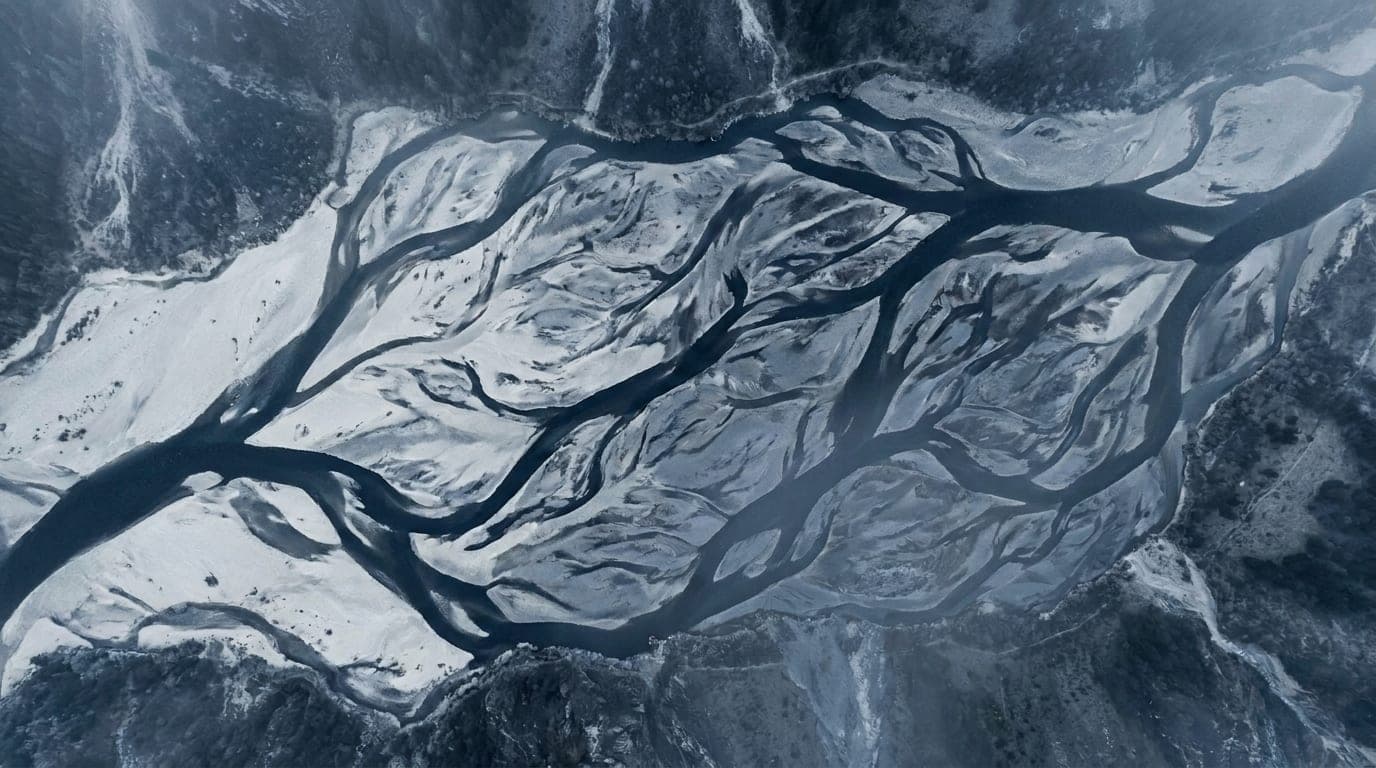 Kühle Alpenlandschaft in Blaustunden-Tönen: ein verzweigter Gletscherfluss aus der Vogelperspektive, dessen silbrige Arme sich über den dunklen Talboden ziehen.