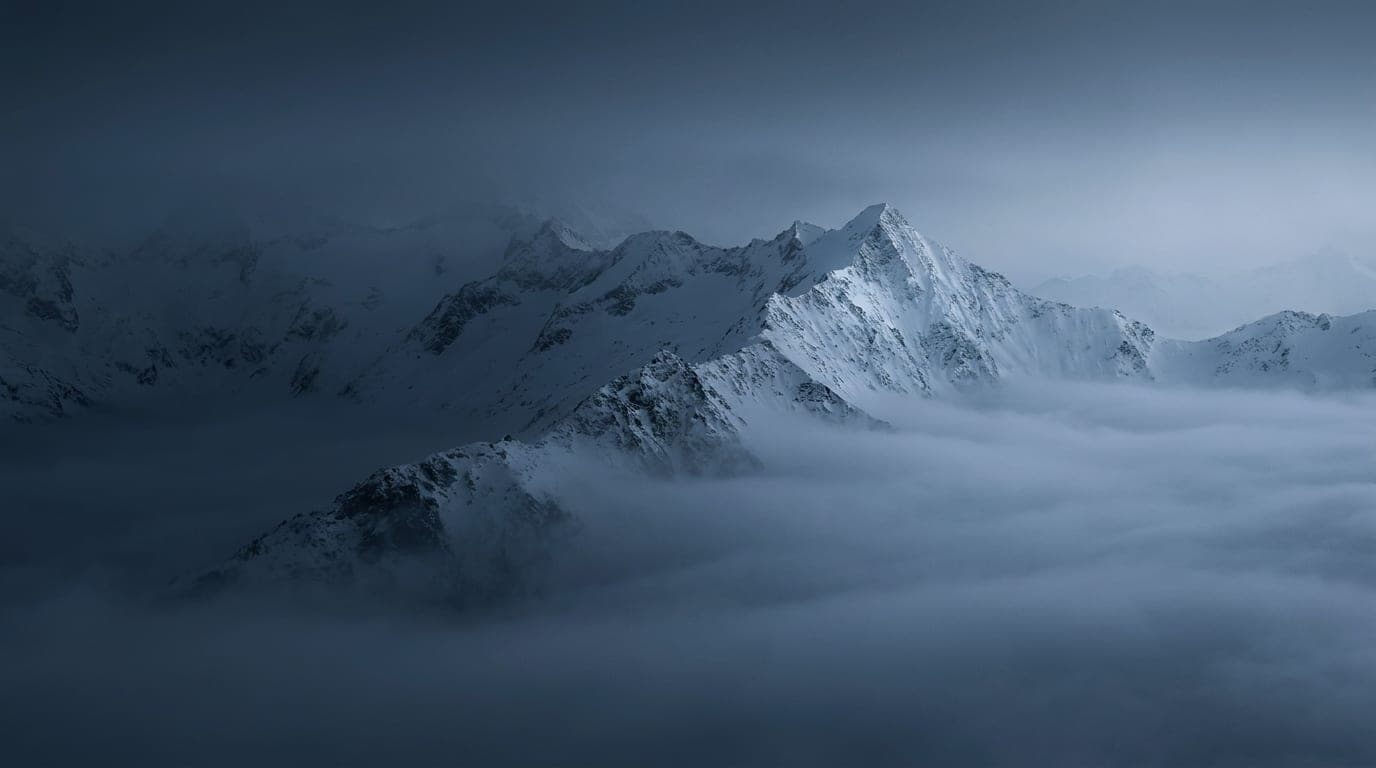 Kühle Alpenlandschaft in Blaustunden-Tönen: ein einzelner scharfer Schneegrat ragt klar über ein Meer aus kaltem Nebel hinaus.