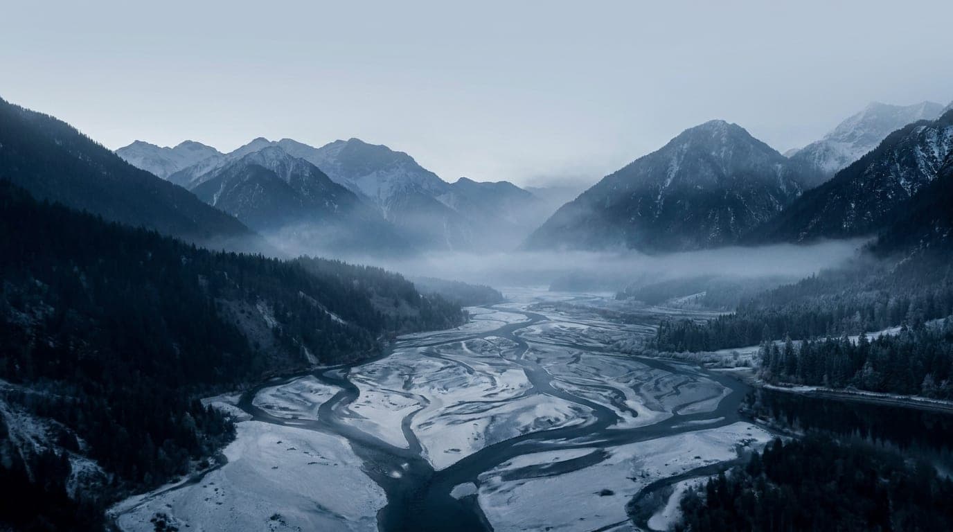 Kühle Alpenlandschaft in Blaustunden-Tönen: ein erhöhter Blick über ein langes Tal, durch das sich ein Fluss zu fernen Gipfeln windet.