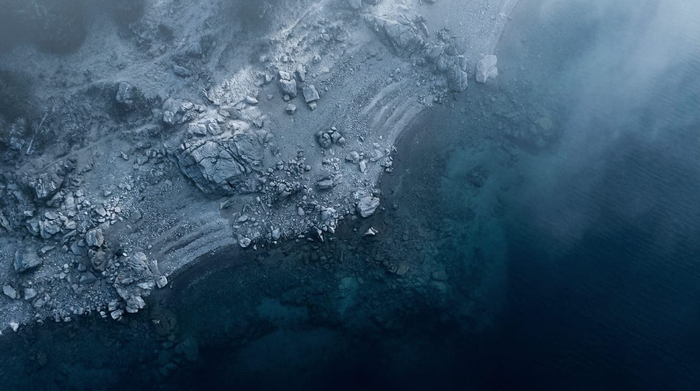 Kühle Alpenlandschaft in Blaustunden-Tönen: ein felsiges Ufer, das aus der Vogelperspektive auf klares kaltes Wasser trifft.