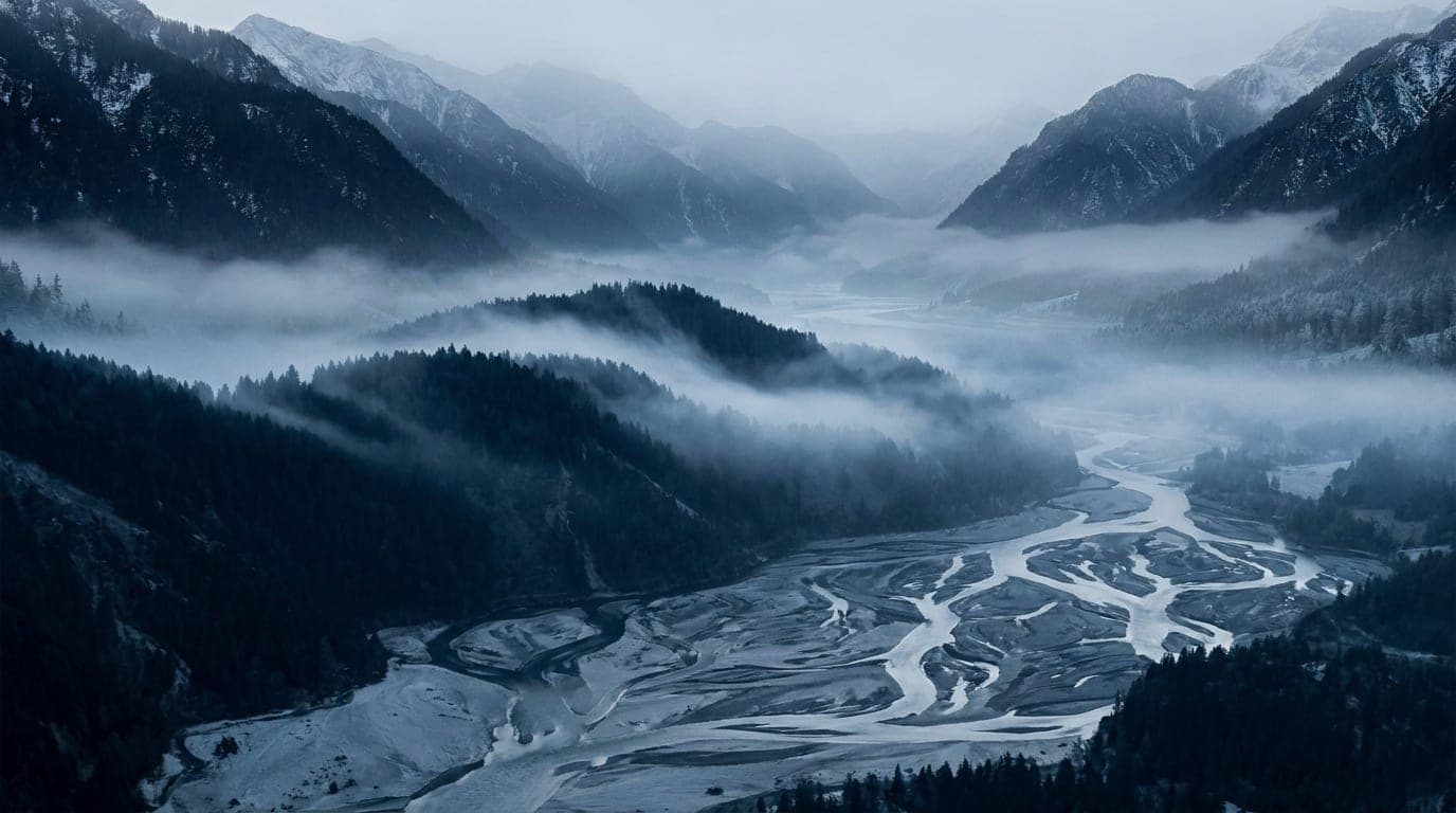 Kühle Alpenlandschaft in Blaustunden-Tönen: kalter Nebel, der zwischen dunkel bewaldeten Bergrücken aufsteigt.